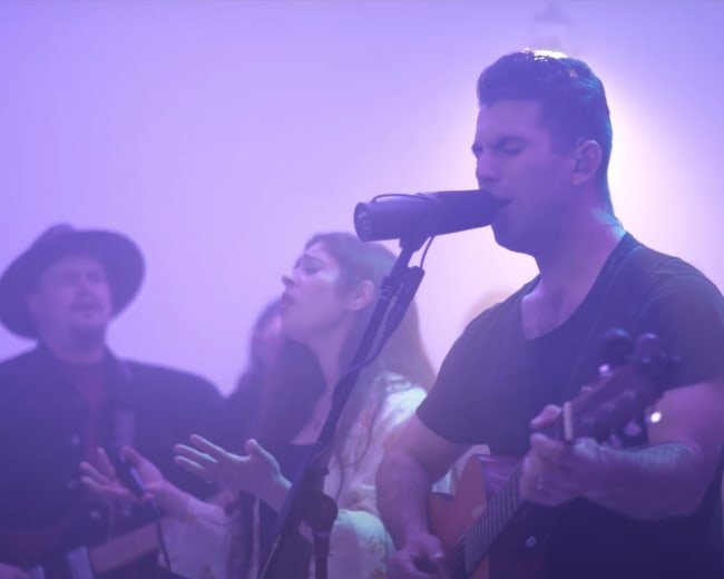 A group of musicians performs on stage with microphone and guitar under a purple light.