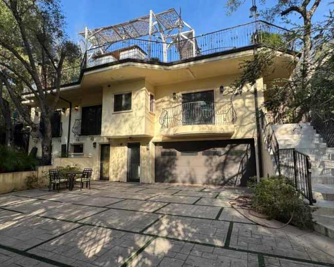 The image depicts a two-story house with a garage, surrounded by trees and featuring a rooftop terrace, along with a patio area with outdoor furniture.