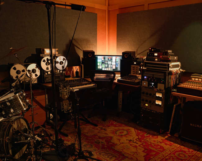 The image shows a music studio with recording equipment, including a drum set, reel-to-reel tape machines, and a computer setup in a soundproofed room.