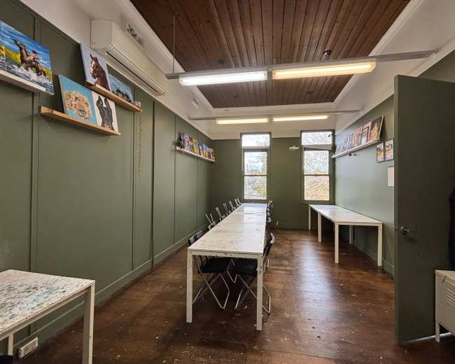 The image shows a spacious room with green walls, wooden flooring, and several tables and chairs arranged around the space, along with artwork displayed on the walls.