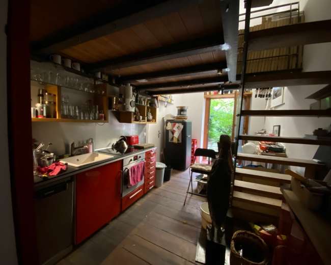 The image shows a compact kitchen with red cabinets, stainless steel appliances, and wooden beams, leading to a stairway and an open doorway filled with greenery.