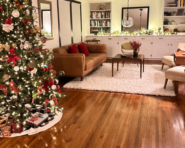 The living room features a decorated Christmas tree, a brown sofa with red cushions, and a coffee table on a plush white rug.