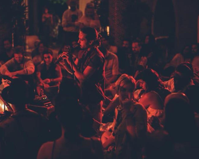 A singer performs onstage while an audience sits closely around, illuminated by soft lights in an outdoor setting.