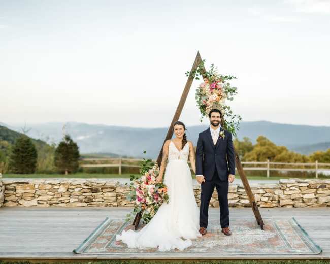 A bride and groom stand in front of a floral-decorated wooden arch with mountains in the background.