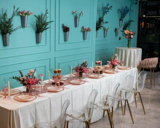 A long dining table is elegantly set with floral arrangements, tableware, and decorative elements against a turquoise wall adorned with potted plants.