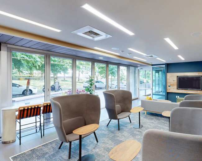 The image shows a modern lounge area with comfortable seating, a television, and large windows that provide natural light.