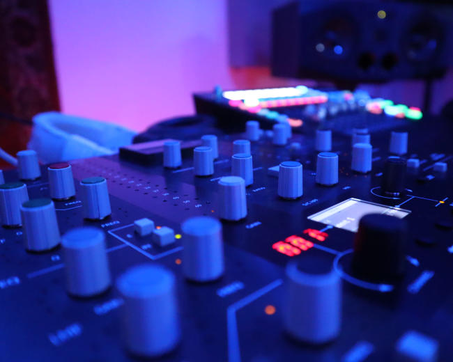 A close-up view of a music production mixer with various knobs and buttons, illuminated by blue and colorful lights in the background.