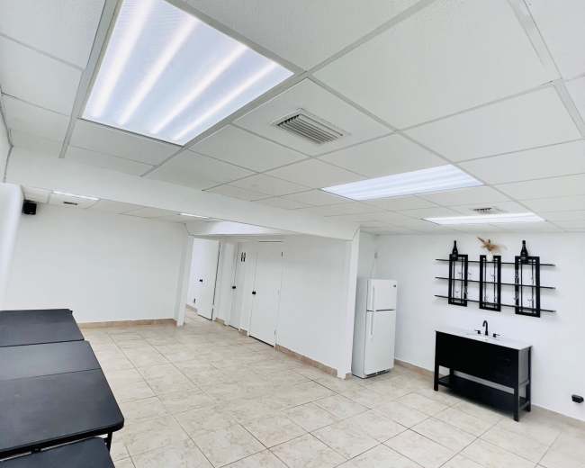 The image shows a spacious room with tiled flooring, fluorescent ceiling lights, a white refrigerator, and a black console table with decorative wall sconces.