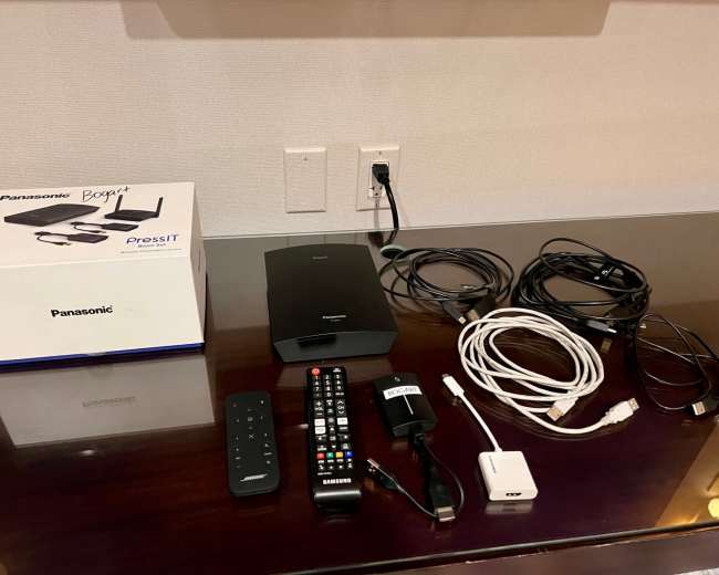 The image shows a Panasonic PressIT box alongside a remote control, various cables, and a power adapter on a table.