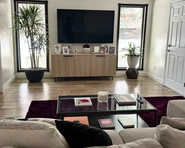 A cozy living room features a large flat-screen TV mounted on a light-colored wall, flanked by windows, with a wooden media console beneath and a coffee table in front of a sectional sofa.