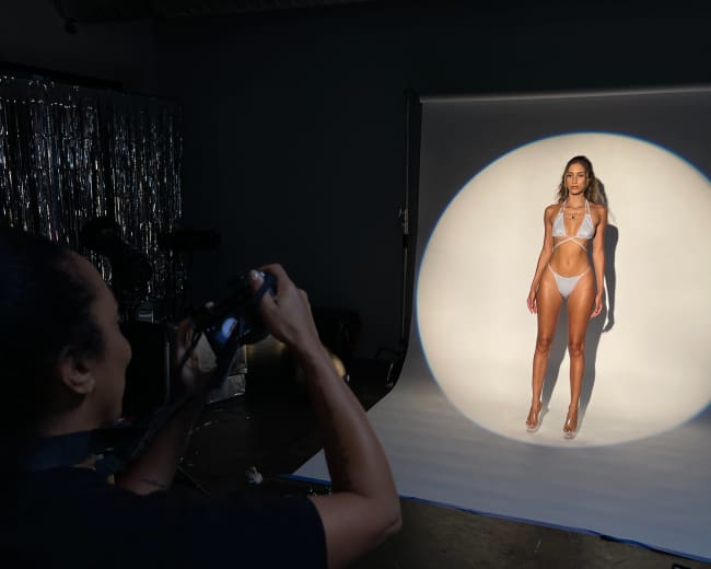 A person uses a camera to photograph a model posing against a lit backdrop in a studio setting.
