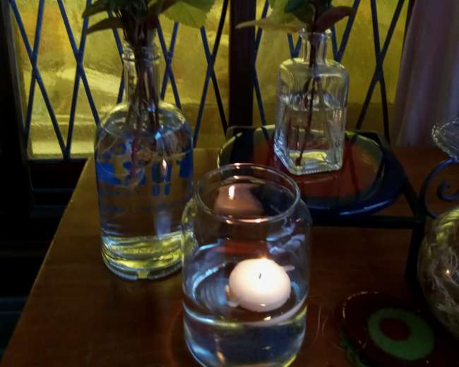 Two vases with greenery and a candle float in a glass jar, situated on a table near a window with stained glass.