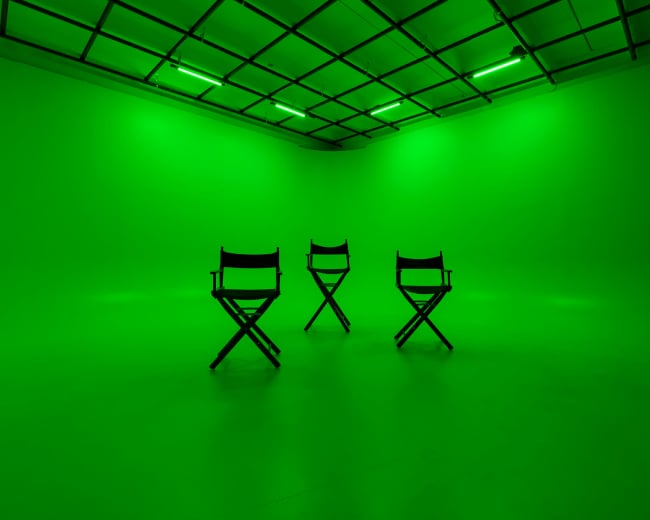 Three empty director's chairs are arranged in a brightly lit green studio space.