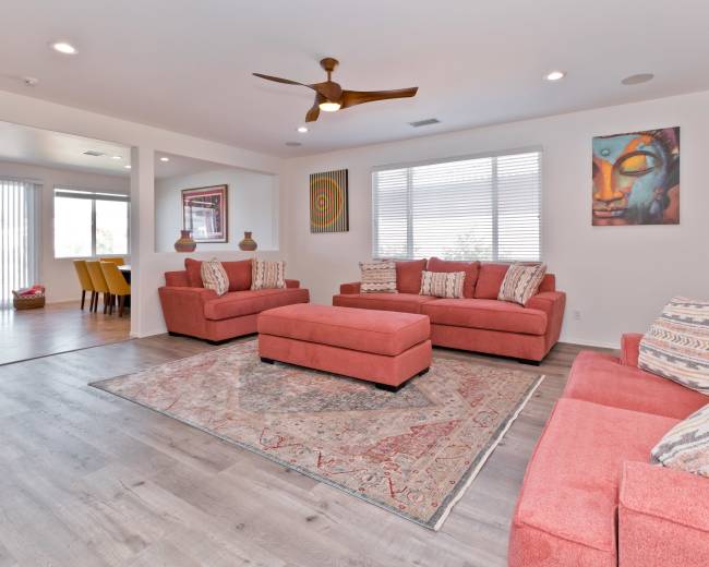The living room features two salmon-colored sofas and an ottoman arranged around a patterned rug, with large windows providing natural light.