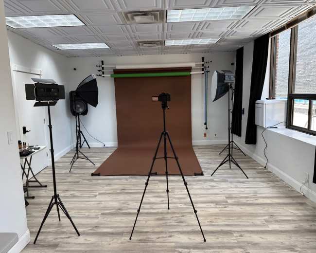 A photography studio with a brown backdrop, two studio lights, a large speaker, and tripods arranged for a photo shoot.
