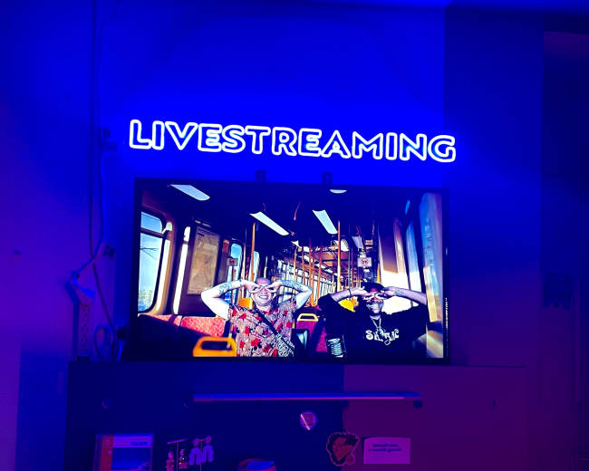 A neon sign reads "LIVESTREAMING" above a television displaying two people in a train car.