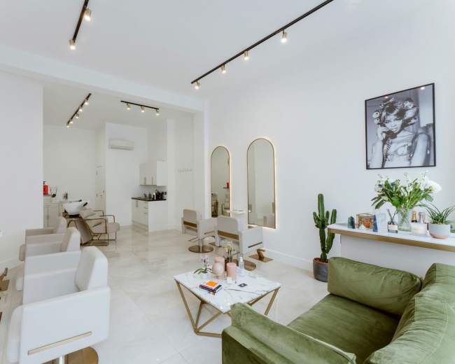 The image shows a modern salon interior with white walls, several styling chairs, mirrors, a coffee table, and decorative plants.