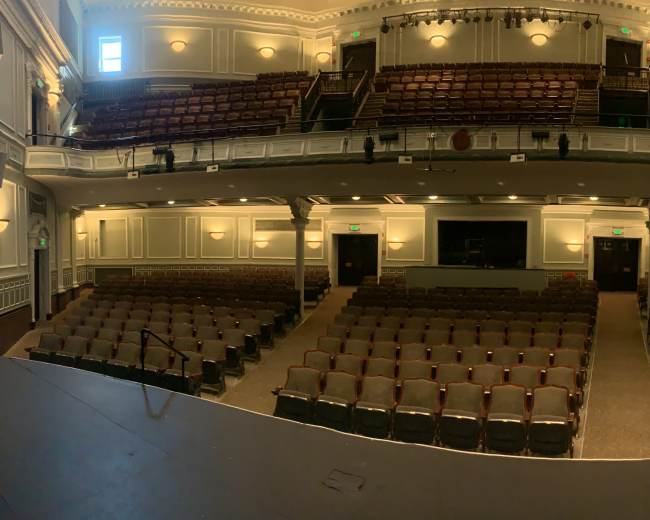 The image shows a nearly empty theater with rows of seats arranged in a semicircle, viewed from the stage.