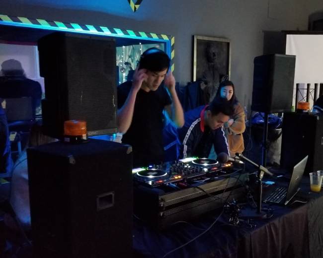Two DJs are performing at a small venue with turntables, speakers, and a laptop, while a person watches in the background.