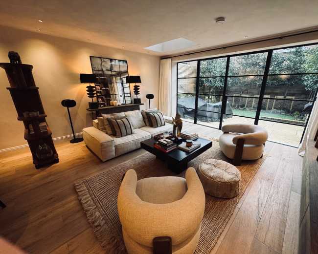 The image shows a modern living room featuring neutral-toned furniture, a large window overlooking a garden, and decorative elements on a coffee table.