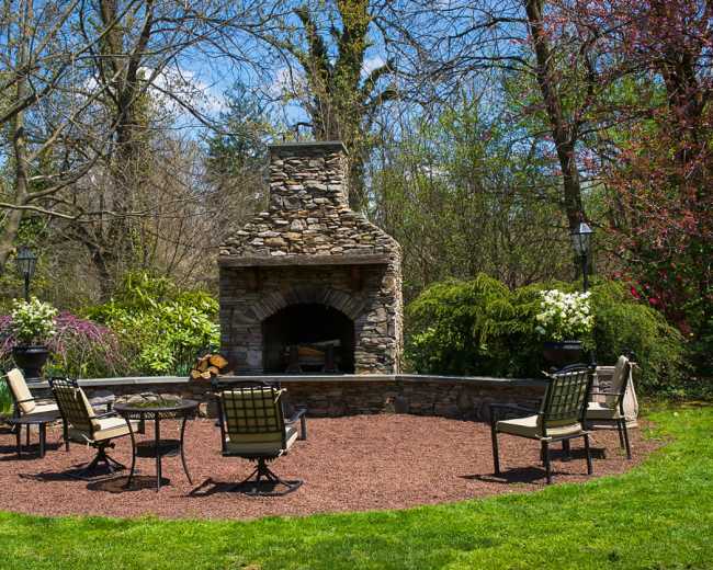 A stone outdoor fireplace is surrounded by several chairs on a gravel patio, with trees and flowering plants in the background.
