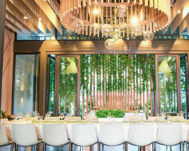 A long rectangular dining table is set in a modern restaurant with large windows revealing greenery outside.
