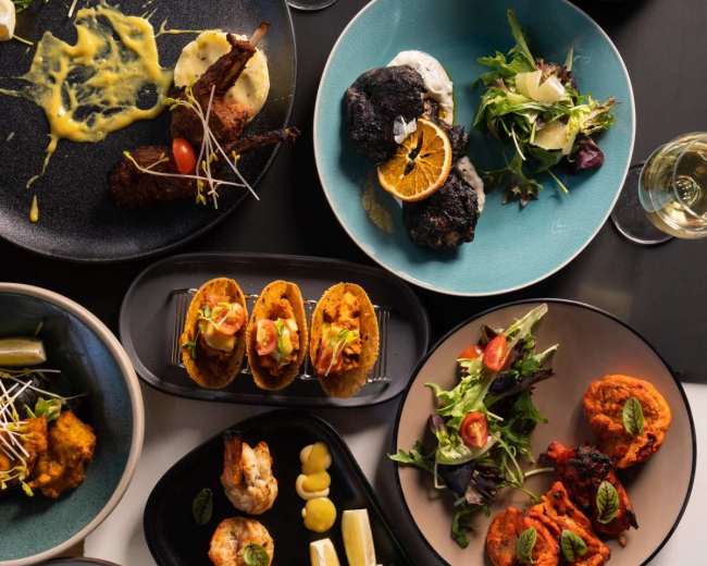 The image displays a variety of dishes arranged on a table, including tacos, grilled shrimp, salads, and a pastry, alongside drinks and sauces.