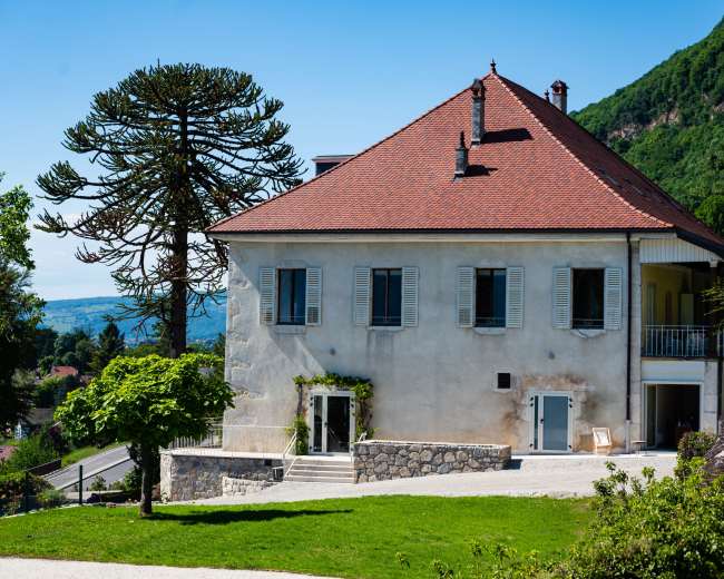 A large two-story house with a red-tiled roof and white walls is surrounded by greenery and a stone pathway, with a prominent tree nearby.