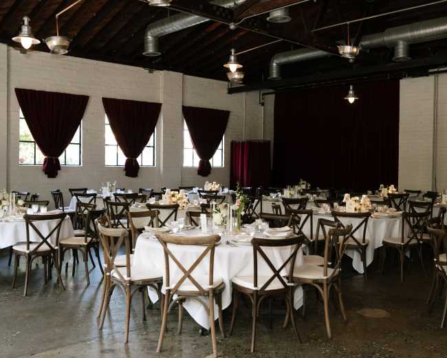The image shows a spacious dining area set with round tables, each adorned with white tablecloths and centerpieces, under wooden beams and red drapery.