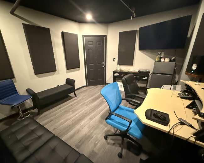The image shows a recording studio featuring a control desk, several chairs, soundproofing panels on the walls, and a closed door.