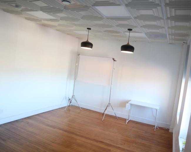 A spacious, empty room with wooden flooring, two pendant lights hanging from the ceiling, and a white backdrop stand near a small white table.