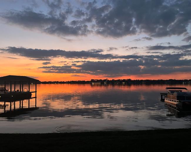 The image shows a tranquil lake at sunset, with a dock on the left and a boat moored on the right, reflecting colorful clouds in the water.
