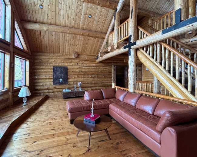 The image shows a spacious log cabin interior featuring a large leather sofa, wooden beams, and expansive windows that allow natural light to fill the room.