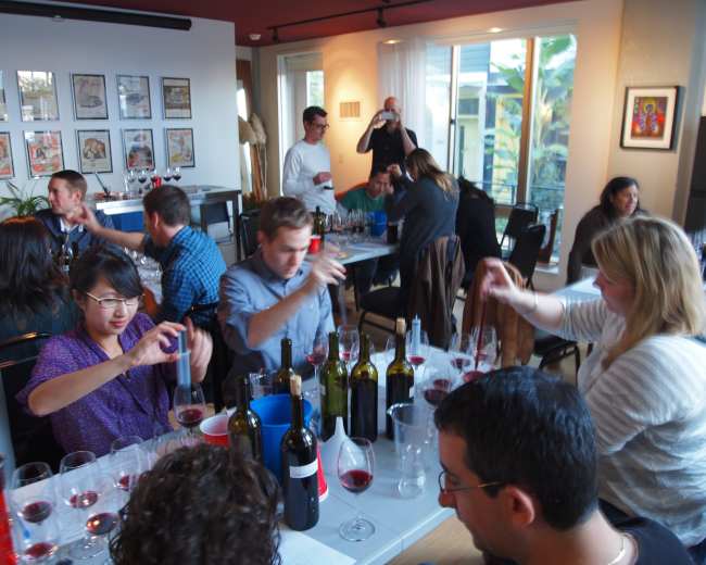A group of people is participating in a wine tasting event around a table filled with glasses and wine bottles in a well-lit room.