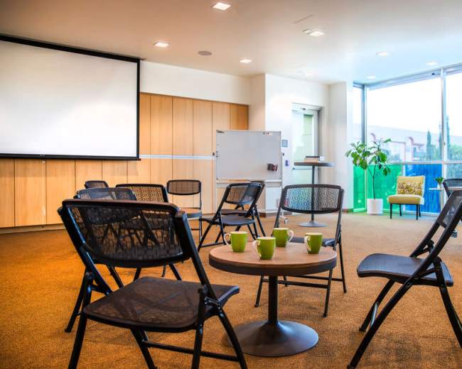 The image shows a meeting room with several chairs arranged around a circular table, a large screen, and windows providing outdoor views.