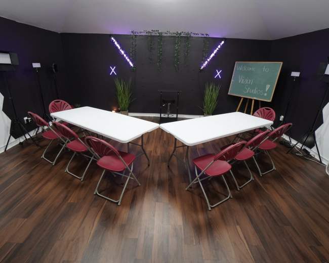 A spacious studio features a large, central arrangement of two tables set in an inverted "V" shape, surrounded by red chairs, with a chalkboard and greenery in the background.