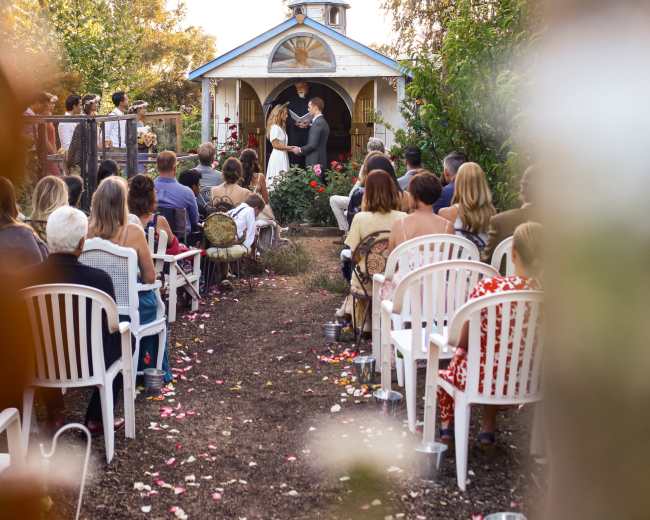 A couple exchanges vows in front of a small chapel while guests sit in white chairs arranged along a path lined with flower petals.