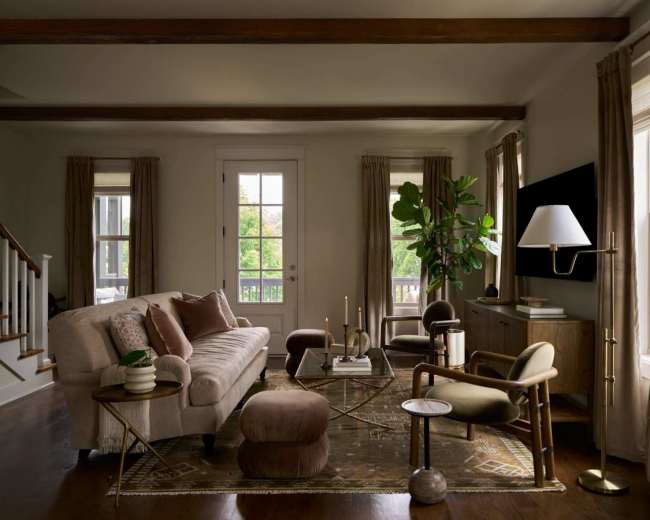 A cozy living room features a beige sofa, a glass coffee table, and large windows with sheer curtains, complemented by plants and decorative accessories.