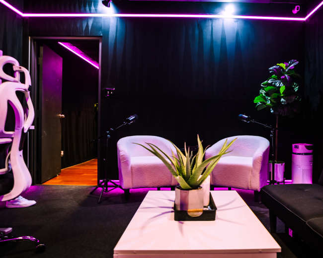 A cozy room features two white chairs, a black futon, a small plant on a coffee table, and a microphone setup under pink neon lighting.