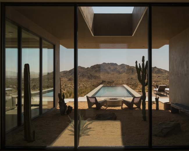 The image shows a modern house with large glass windows framing a pool and desert landscape, featuring cacti and mountains in the background.