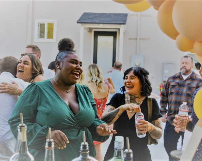 A group of friends enjoys drinks and laughter at a festive outdoor gathering near a bar setup with balloons in the background.