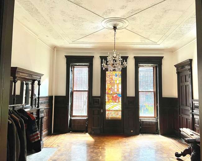 The image shows a spacious, empty room featuring dark wooden trim, large windows with stained glass, a chandelier, and wooden furniture along the walls.