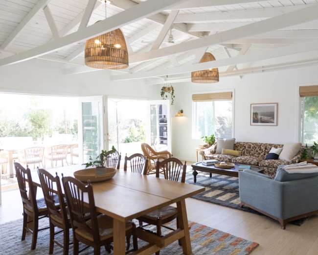A bright, open living space features a dining table with wooden chairs, a comfortable sofa, and large windows allowing natural light to fill the room.