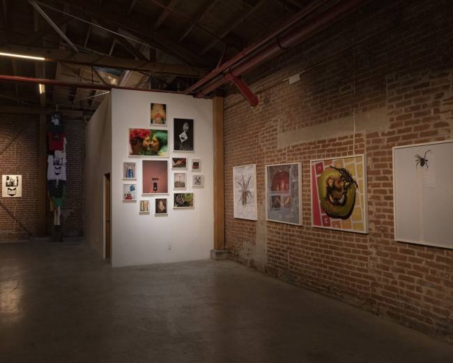 The image shows an art gallery with brick walls displaying various framed artworks, including photographs and illustrations, in a spacious, industrial-style setting.