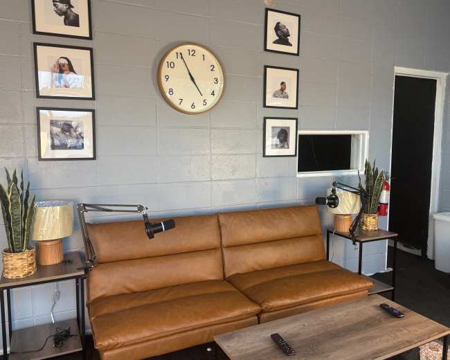 The image shows a modern living room with a brown leather couch, two lamps, a clock on the wall, and framed photographs.