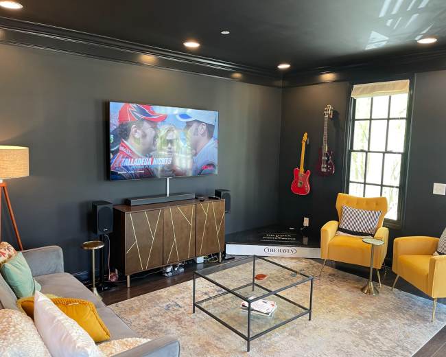 A modern living room features a large television mounted on a black wall, with a stylish gray sofa, yellow chairs, and a glass coffee table, along with decorative items and a window.