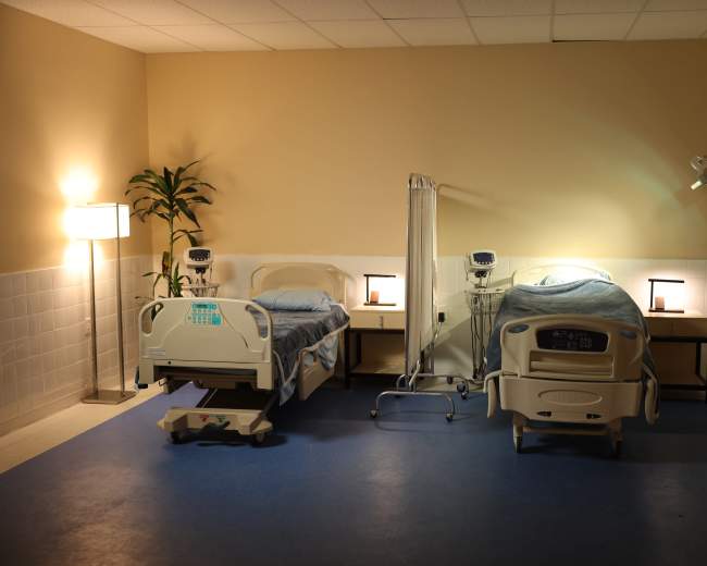 The image shows a hospital room with two beds, a lamp, medical equipment, and a plant against the wall.