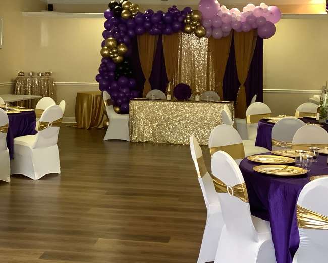 The image shows a banquet hall decorated with purple, gold, and white balloons and table settings for an event.