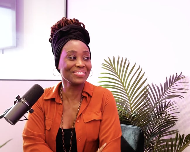 A woman with a head wrap and orange shirt sits in front of a microphone and a plant.