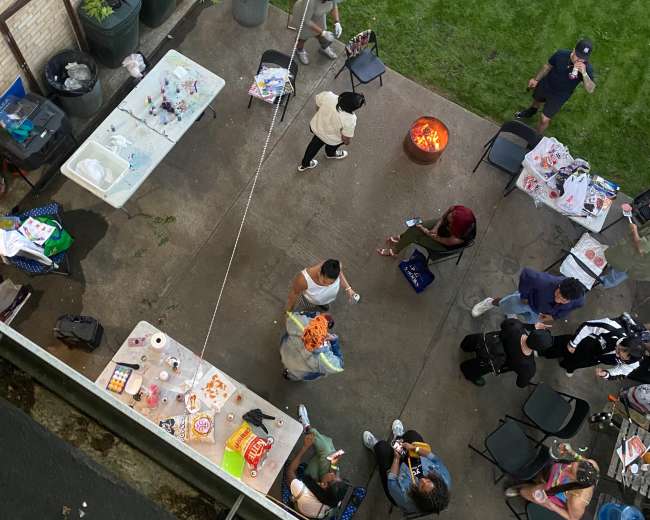 A group of people gathers in a backyard, with tables set up and a fire pit in use.